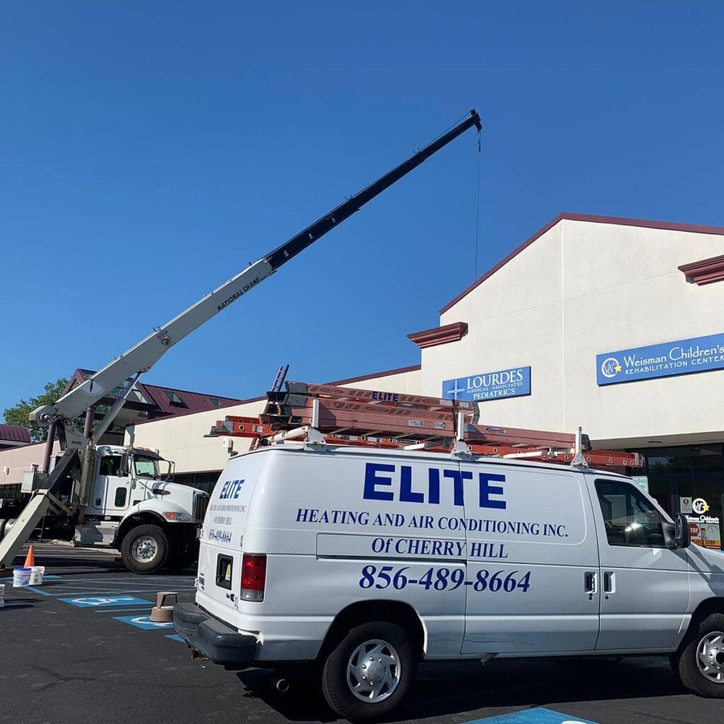 A crane lifts equipment onto the roof of a building while a white Elite Heating and Air Conditioning van, proudly displaying our Careers sign, is parked in the lot below.