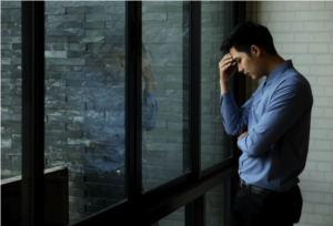 A man in a blue shirt stands by a window with his hand on his forehead, looking down with a thoughtful expression—perhaps worried about common failures in commercial HVAC systems and their prevention.