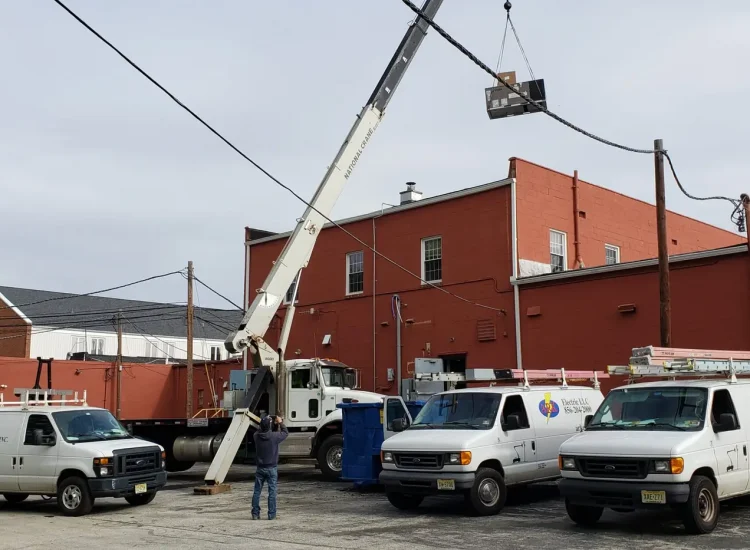 Three white utility vans are parked in a lot near a red brick building, showcasing Elite's contact details. A crane lifts an air conditioning unit onto the roof for a commercial HVAC repair, as a person guides the operation. An overcast sky and power lines frame this scene of expertise.