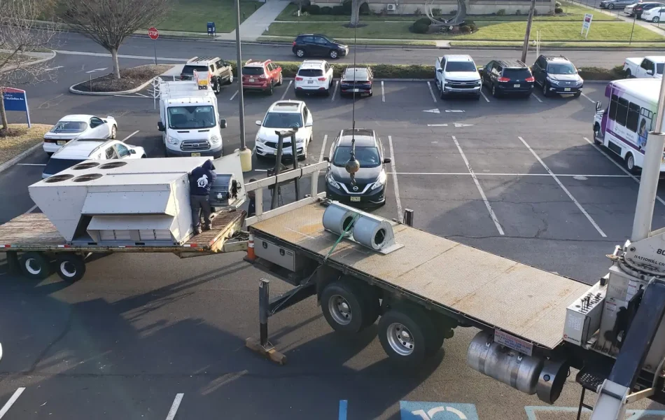 A flatbed truck loaded with large HVAC units is parked in a lot where a worker from one of the best HVAC companies near you is adjusting the units. Nearby are several cars and a delivery truck. In the background, a purple and white bus stands beside a leafless tree and building under the clear sky.