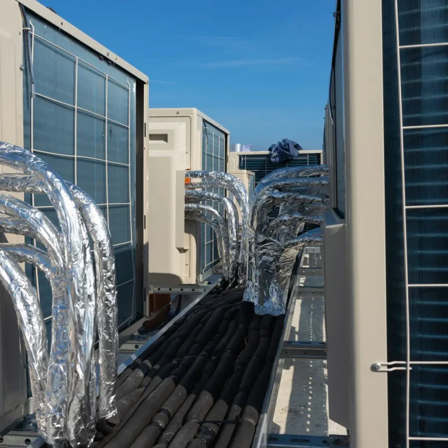 A rooftop view showcases the impressive setup of commercial air conditioning units, their interconnected pipes wrapped in shiny silver foil. Mounted on a sturdy metal platform under a clear blue sky, this scene reflects the expertise of top-tier Commercial HVAC repair services.