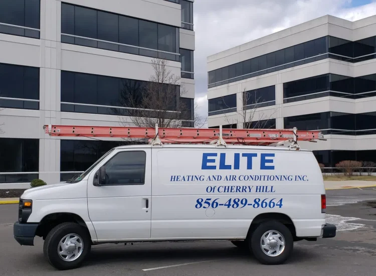 A white van with Elite Heating and Air Conditioning Inc. of Cherry Hill and a phone number is parked in front of modern office buildings. A red ladder is secured to the roof, ready for commercial HVAC repair. The sky is partly cloudy, with leafless trees lining the paved driveway.